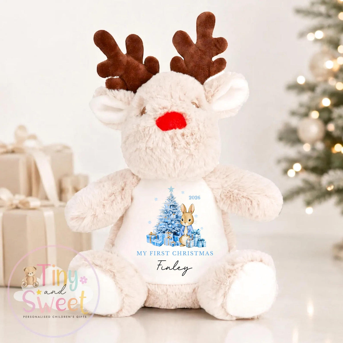 May include: A plush reindeer toy with soft beige fur, brown antlers, and a bright red nose. The chest features a printed graphic of a blue Christmas tree, presents, a rabbit, the text "MY FIRST CHRISTMAS", the year "2026", and the name "Finley".
