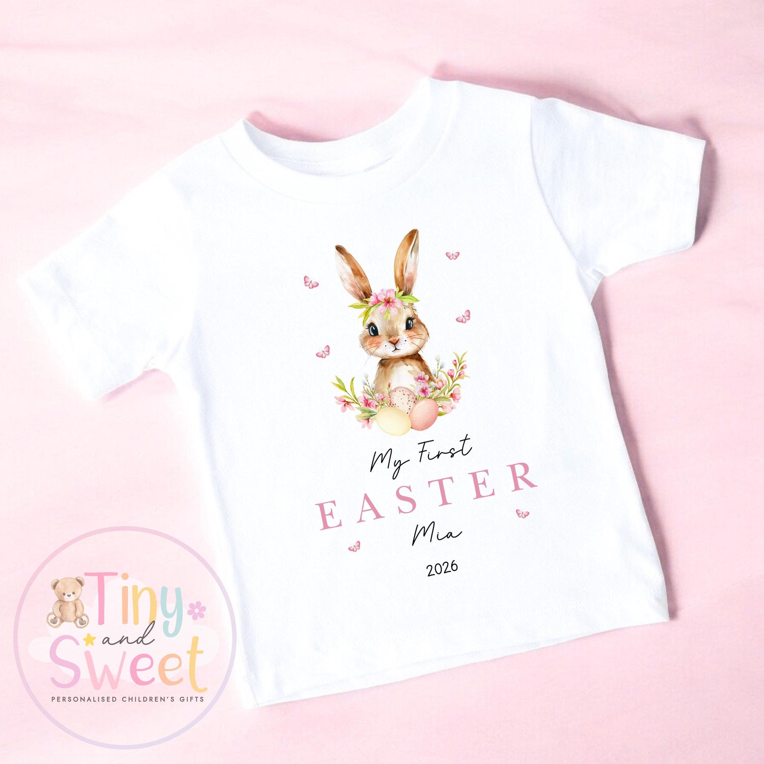 May include: White t-shirt with a watercolour illustration of a bunny wearing a floral crown, surrounded by flowers and butterflies. The text &amp;quot;My First Easter Mia 2026&amp;quot; is printed in pink. A personalised children&amp;