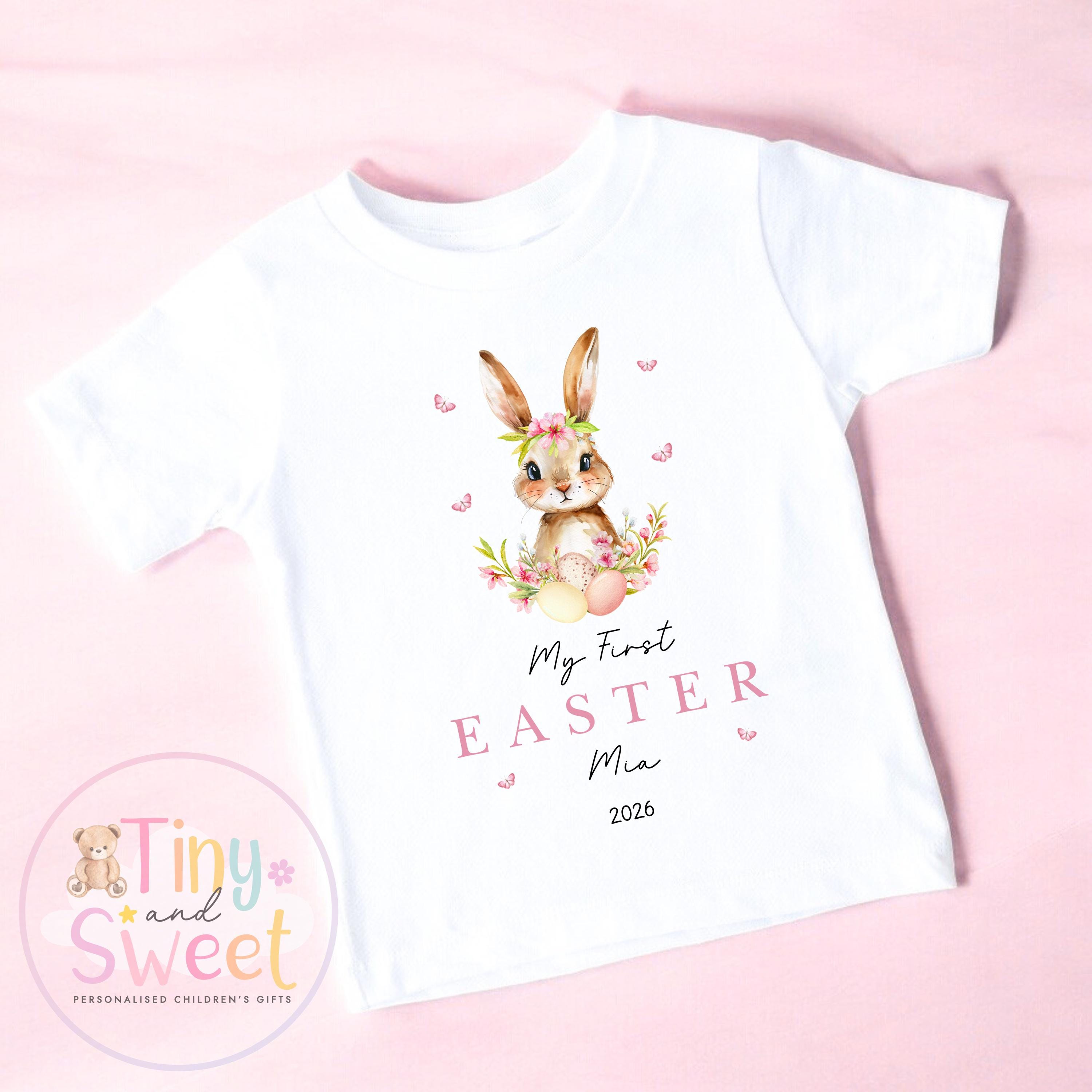 May include: White t-shirt with a watercolour illustration of a bunny wearing a floral crown, surrounded by flowers and butterflies. The text &amp;quot;My First Easter Mia 2026&amp;quot; is printed in pink. A personalised children&amp;