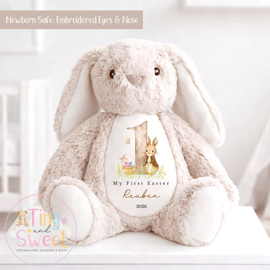Beige plush bunny teddy sitting on a white surface with embroidered eyes and nose, featuring a “My First Easter” design on its belly with the number 1, a bunny illustration, and personalized text reading “Reuben 2026.”