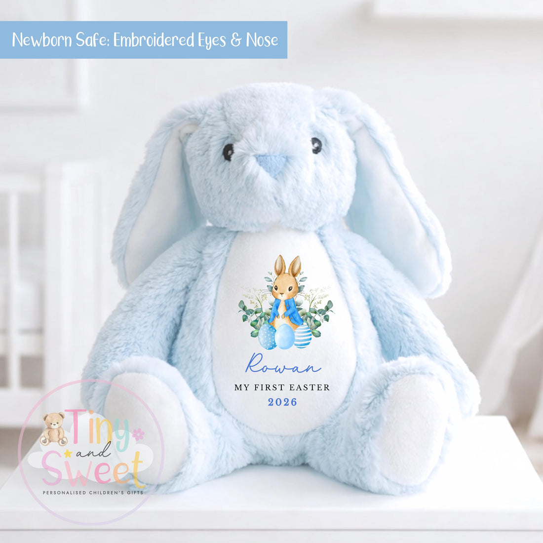 Light blue plush bunny teddy sitting on a white surface with embroidered eyes and nose, featuring a “My First Easter” design on its belly with a bunny illustration, blue Easter eggs, and personalized text reading “Rowan 2026.”