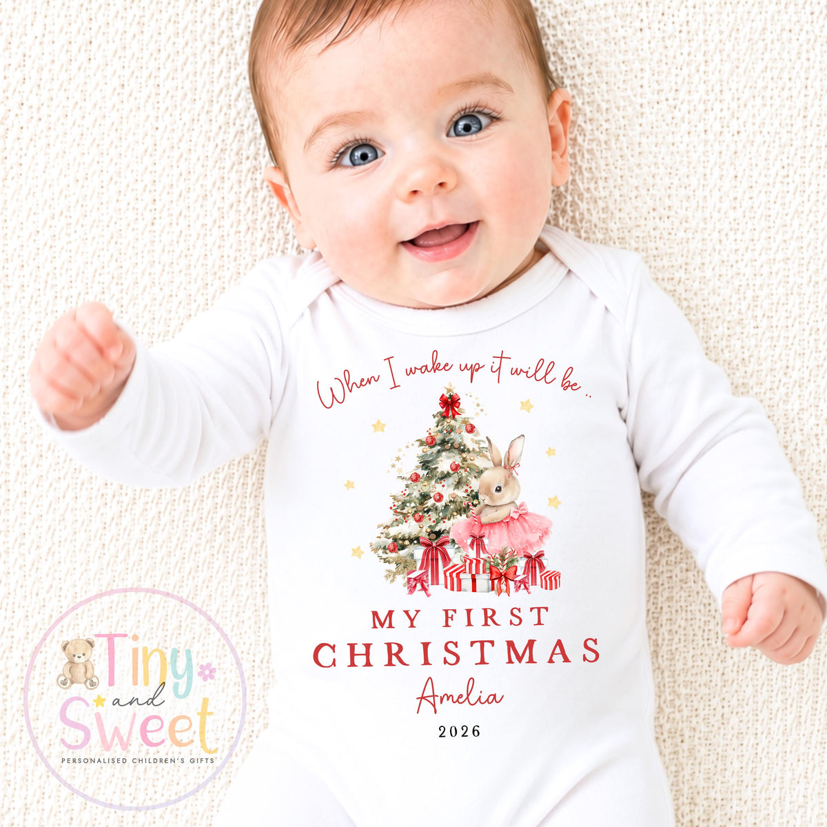 May include: A white long-sleeve baby bodysuit featuring a graphic of a Christmas tree, a rabbit in a pink tutu, and wrapped gifts. The text reads: When I wake up it will be.. MY FIRST CHRISTMAS Amelia 2026. The design includes small gold stars.